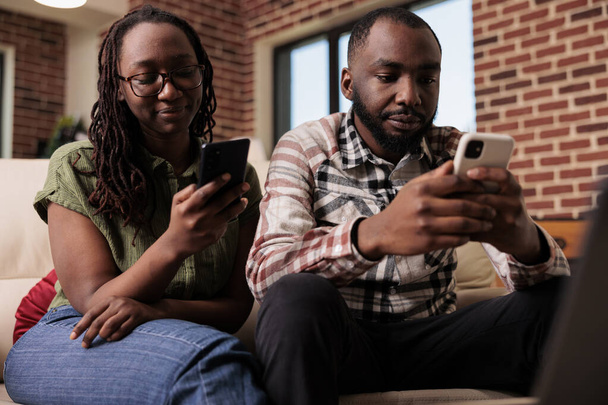 アフリカ系アメリカ人のカップルは、ソーシャルメディアのコンテンツをスマートフォンで見て、ソファでお互いを無視することに時間を費やしています。男性と女性は携帯電話を使った議論の後一緒に動揺して住んでいます. - 写真・画像
