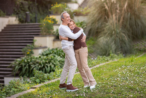 Senior Life Happiness. Starší manželé objímající stojící v zeleném parku venku, pózující hledá Aside. Milující manžel objímání manželka během procházky v zahradě města. Full Length Shot - Fotografie, Obrázek