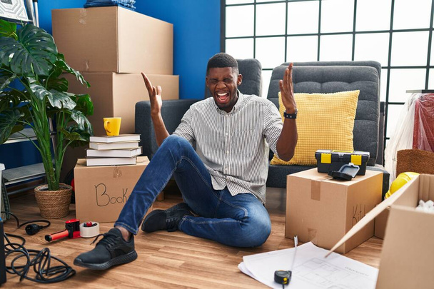 Afroamerikaner, der in seinem neuen Zuhause auf dem Boden sitzt und mit erhobenen Armen und geschlossenen Augen aufgeregt den Erfolg feiert. Siegerkonzept  - Foto, Bild