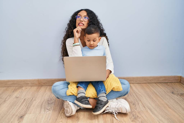 床に座っているコンピュータのラップトップを使用して若いヒスパニック系の母親と子供は、質問を心配し、心配し、顎に手で緊張  - 写真・画像