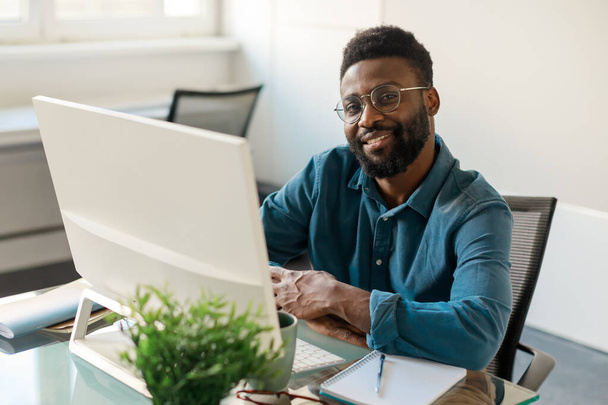 Positive black businessman in eyeglasses smiling at camera, sitting in front of computer at workdesk in office interior, copy space. Career, business leader - 写真・画像