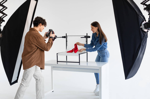 Joven fotógrafo masculino tomando fotos de elegantes zapatos rojos en el fondo de la plataforma blanca en el fotoestudio con equipos modernos, señora ayudando a hacer sesión de fotos de contenido - Foto, imagen