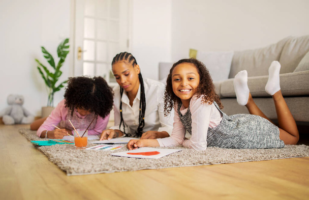 週末に家で一緒に時間を過ごすお母さんと妹と一緒に描く陽気なアフリカ系アメリカ人の子供の女の子,床に横になってカメラに笑顔を掲載.家族趣味。選択的フォーカス - 写真・画像