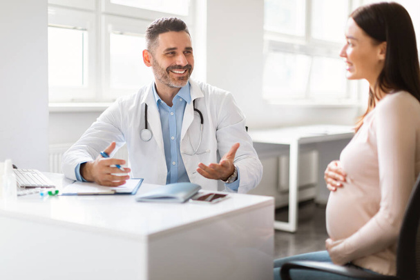 Pregnant woman visiting doctor man at clinic, therapist giving recommendations to happy expecting lady. Healthcare during pregnancy concept - Фото, зображення