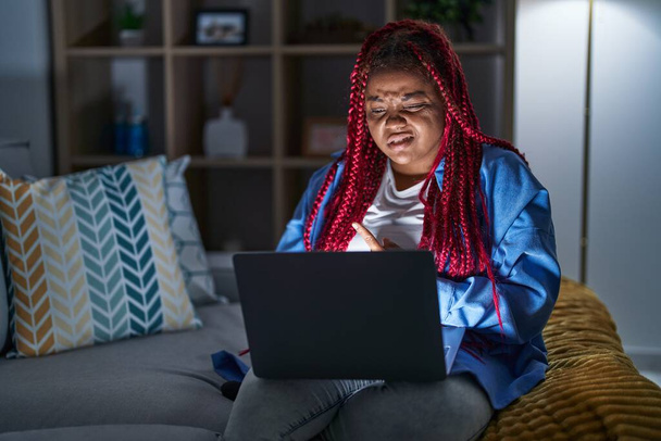 Mujer afroamericana con cabello trenzado usando computadora portátil en la noche señalando a un lado preocupado y nervioso con el dedo índice, expresión preocupada y sorprendida  - Foto, imagen
