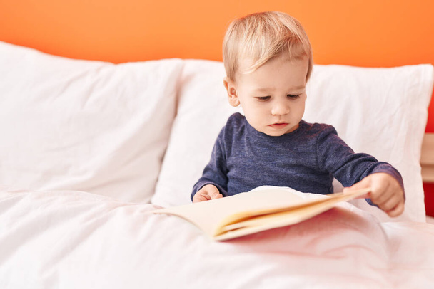 Adorable blond toddler reading story book sitting on bed at bedroom - Zdjęcie, obraz
