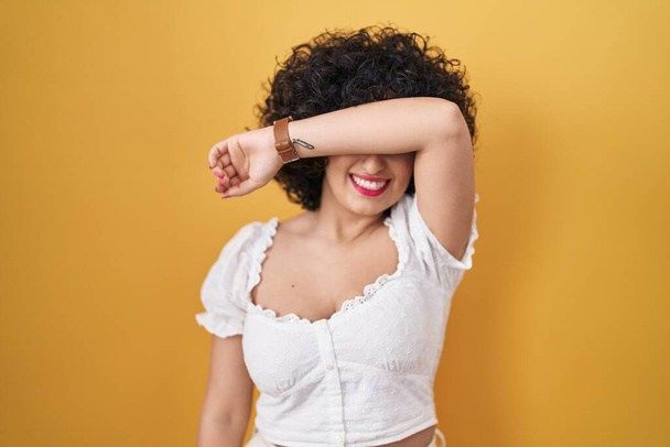 Young brunette woman with curly hair standing over yellow background covering eyes with arm smiling cheerful and funny. blind concept.  - Fotó, kép
