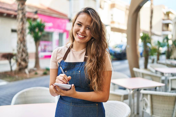 Young beautiful hispanic woman waitress smiling confident writing on notebook at coffee shop terrace - Фото, изображение