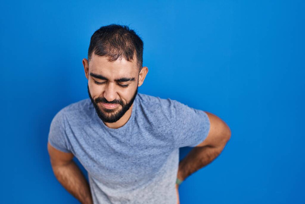 Middle east man with beard standing over blue background suffering of backache, touching back with hand, muscular pain  - Фото, зображення