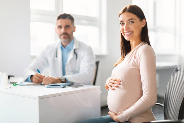 Happy young pregnant woman sitting at clinic during consultation with middle aged male gynecologist, embracing belly and smiling at camera, selective focus on lady - Fotografie, Obrázek
