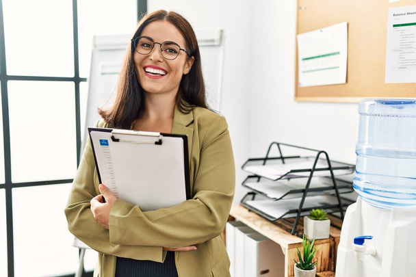 Νέα όμορφη ισπανόφωνη εργαζόμενη επιχειρηματίας χαμογελά αυτοπεποίθηση κρατώντας πρόχειρο στο γραφείο - Φωτογραφία, εικόνα