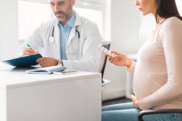 Léky v těhotenství. Lékař předepisující léky pro mladou těhotnou dámu, pacientka držící blistr s tabletami, sedící na klinice, selektivní zaměření - Fotografie, Obrázek