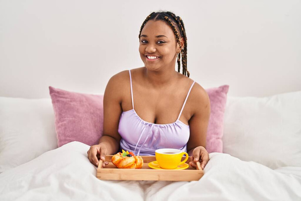 Αφροαμερικανή γυναίκα με πλεξούδες κρατώντας δίσκο με πρωινό φαγητό στο κρεβάτι φαίνεται θετική και ευτυχισμένη στέκεται και χαμογελά με αυτοπεποίθηση χαμόγελο που δείχνει τα δόντια  - Φωτογραφία, εικόνα