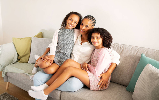 Happy Black Mother And Kid Daughters Cuddling Embracing Sitting On Sofa At Home, Smiling Looking At Camera. Shot Of Family Of Three Bonding In Modern Living Room On Weekend - Photo, image