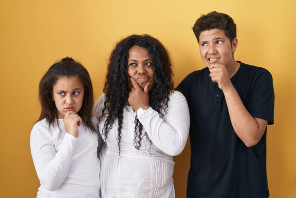 Family of mother, daughter and son standing over yellow background thinking worried about a question, concerned and nervous with hand on chin  - Fotoğraf, Görsel