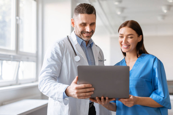 Happy male doctor and female nurse talking, working together, using laptop, discussing patient medical checkup results or diagnosis, wearing uniform holding electronic device - Photo, Image
