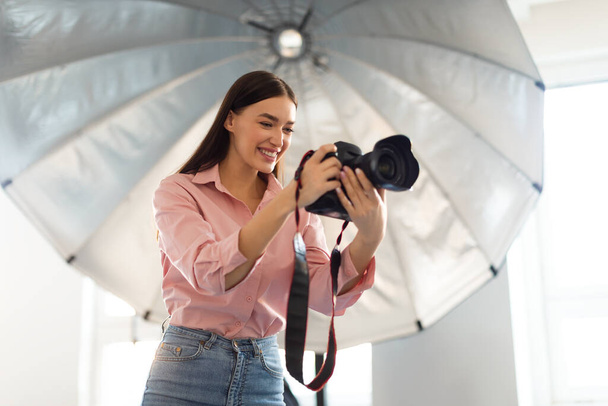 Young female photographer standing in front of reflective umbrella, working with professional dslr camera in modern photostudio, free space - Foto, afbeelding