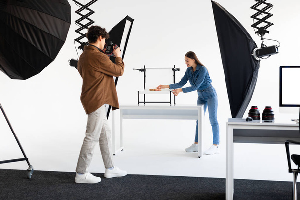 Jovem fotógrafo masculino com fotocâmera tirando fotos de composição na plataforma branca, enquanto sua assistente senhora ajudá-lo no photostudio - Foto, Imagem
