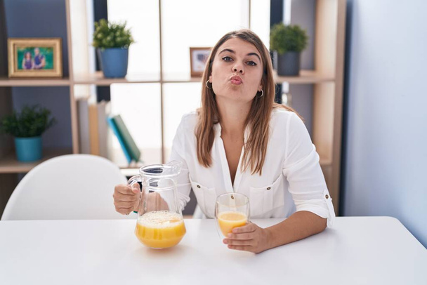 Young hispanic woman drinking glass of orange juice looking at the camera blowing a kiss being lovely and sexy. love expression.  - Foto, Imagem