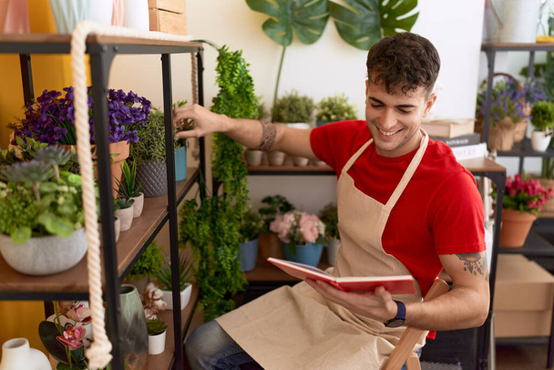 Young hispanic man florist reading book holding plant of shelving at flower shop - Photo, Image