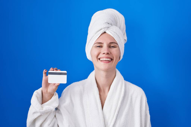 Blondi valkoihoinen nainen yllään kylpytakki tilalla luottokortti näyttää positiiviselta ja onnellinen seisoo ja hymyilee luottavainen hymy osoittaa hampaat  - Valokuva, kuva