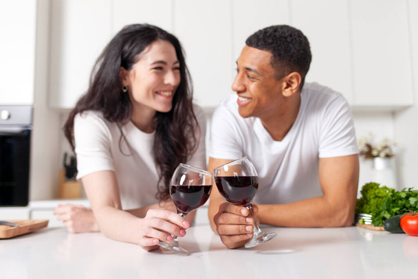 молодая межрасовая пара, пьющая красное вино на кухне и разговаривающая, афроамериканец, звенящий в очках с девушкой и флиртующий, женатая пара, держащая бокалы вина и улыбающаяся - Фото, изображение
