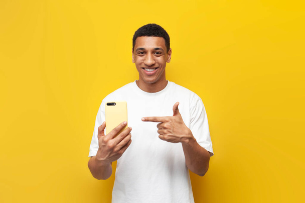 giovane ragazzo afroamericano in t-shirt bianca punta il dito sullo smartphone su sfondo giallo isolato, l'uomo pubblicizza e raccomanda il telefono - Foto, immagini