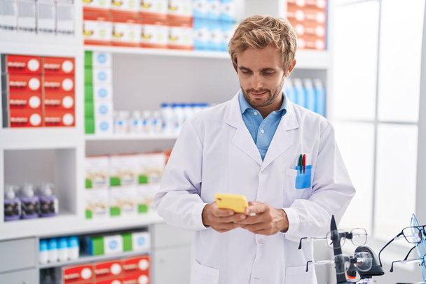 Young man pharmacist using smartphone working at pharmacy - Zdjęcie, obraz