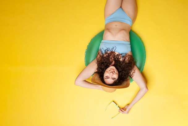 young girl in blue swimsuit lies on inflatable swimming circle on yellow background, woman in sunglasses in the summer on the beach sunbathes, top view - Foto, Imagem