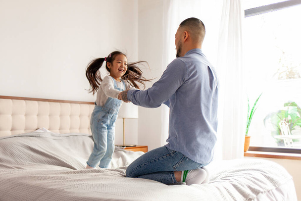 small active Asian girl jumps and dances on the bed at home with her dad, Korean man plays with his daughter and holds her hands, parent raises a child - Foto, Bild