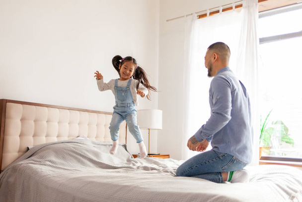 kis aktív ázsiai lány játszik az apjával otthon, energikus gyermek ugrál és táncol az ágyon a szülővel, koreai férfi feldobja a lányát, és örül - Fotó, kép