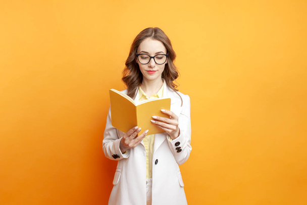 jeune fille étudiante en lunettes et costume blanc lit livre jaune et sur fond isolé de couleur, femme enseigne des leçons - Photo, image
