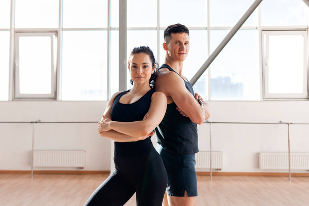 beautiful athletic couple in sportswear stands in the gym in morning, fitness girl and coach man stand back to back in training in bright room - Fotografie, Obrázek