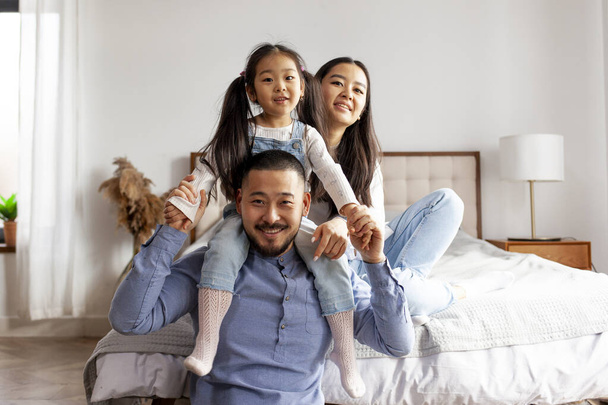 happy asian family sit together at home in the bedroom and smile, little korean girl sits on the shoulders of her dad and rejoices with her parents - Photo, image