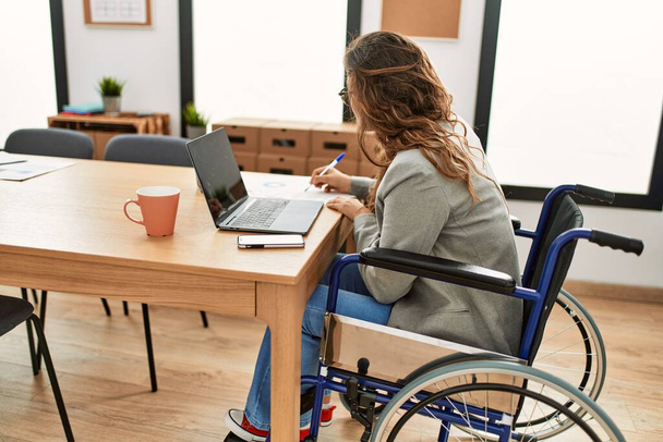 Νεαρή όμορφη ισπανόφωνη επιχειρηματίας που γράφει σε έγγραφο που κάθεται σε αναπηρικό καροτσάκι στο γραφείο - Φωτογραφία, εικόνα