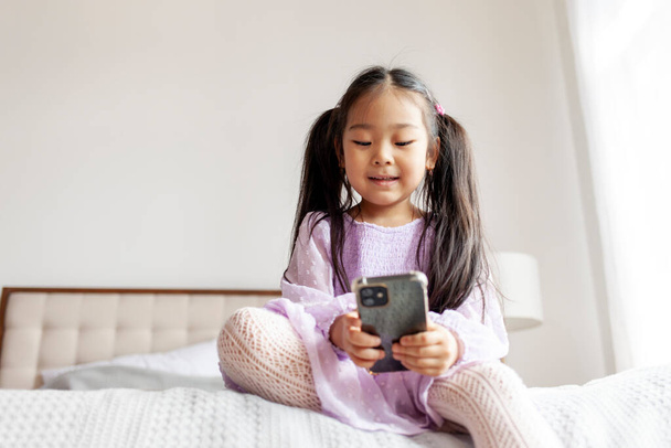 小さなアジア系の女の子が家のベッドに座ってスマートフォンを使って子供は電話を見てメッセージを入力して紫色のドレスで笑っています - 写真・画像