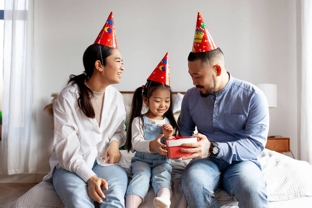 glückliche asiatische Familie in festlicher Kleidung feiert den Geburtstag ihrer kleinen Tochter und schenkt ihr, koreanisches Mädchen feiert Geburtstag mit den Eltern zu Hause, elterliche Liebe und Fürsorge - Foto, Bild