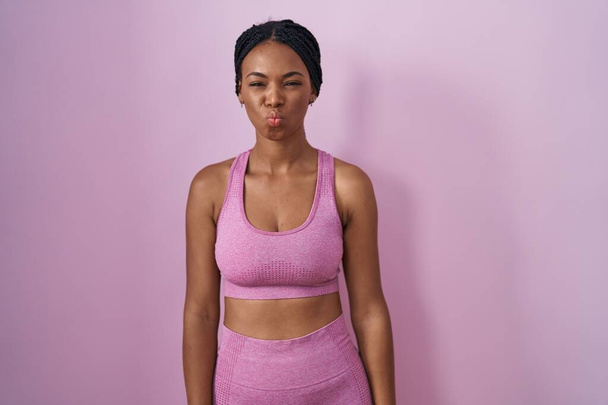 ピンクの背景にスポーツウェアを着たアフリカ系アメリカ人女性が面白い顔で頬をむいています。口は空気で膨らみ狂気の表情で.  - 写真・画像