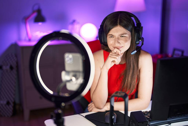 Młoda kaukaska kobieta gra w gry wideo nagrywając ze smartfonem poważną minę myśląc o pytaniu z ręką na podbródku, myśląc o mylącym pomyśle  - Zdjęcie, obraz