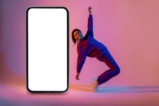 Activa bailarina afroamericana de hip-hop bailando cerca de un enorme smartphone con pantalla en blanco sobre fondo de neón rosa, espacio libre, maqueta. Aplicación de danza o sitio web - Foto, Imagen