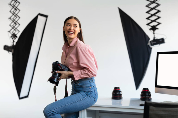 Excited female photographer sitting on edge of table, holding her DSLR camera, looking aside and smiling, working with computer in modern photostudio interior - Фото, зображення