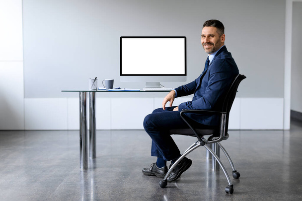 Happy middle aged businessman sitting on desk with computer with empty blank screen at office, free space, mockup template. People, technology, business work concept - Photo, Image