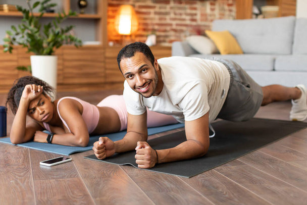 Glad young black guy and tired sad lady in sportswear rest on floor from workout together in living room interior. Huge work with sport app, body care remote, weight loss, yoga and fitness at home - Photo, Image