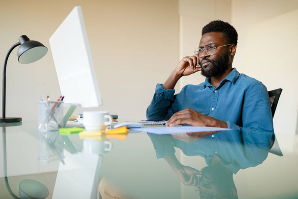 Сконцентрированный черный человек в очках, работающий за компьютером в офисе, проверяющий проект, решающий повседневные вопросы или имеющий видеовстречу, сидящий перед монитором, копирующий пространство - Фото, изображение