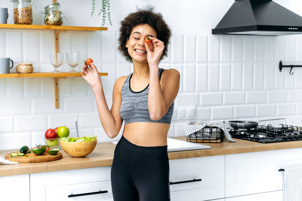 Healthy food, diet. Joyful pretty african american young woman with curly hair in a sportswear, stands in the kitchen, prepares a salad, have fun covering one her eye with tomato, smiles - Photo, image