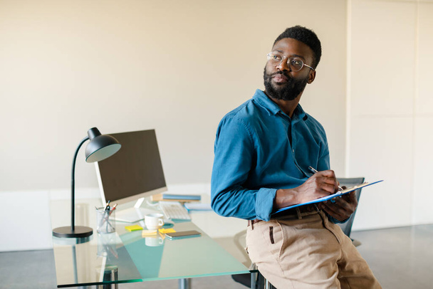 Portret troskliwego czarnego mężczyzny stojącego w biurze koło miejsca pracy, trzymającego schowek i patrzącego na wolną przestrzeń, myślącego o projekcie biznesowym lub problemach - Zdjęcie, obraz