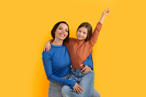 Ευτυχισμένη νεαρή μητέρα και η μικρή κόρη αγκαλιάζοντας και διασκεδάζοντας μαζί, αγαπώντας την οικογένεια των δύο μαμά και προεφηβική θηλυκό παιδί ποζάρει απομονωμένη πάνω από κίτρινο φόντο στο στούντιο, αντιγραφή χώρο - Φωτογραφία, εικόνα