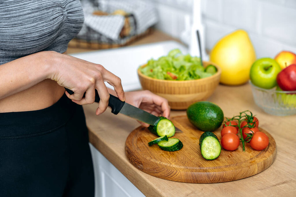 Healthy eating concept. Close-up hands of african american young woman in a sportswear, standing at home in kitchen cutting cucumber while preparing fresh useful salad, smiling happily - Foto, Imagem