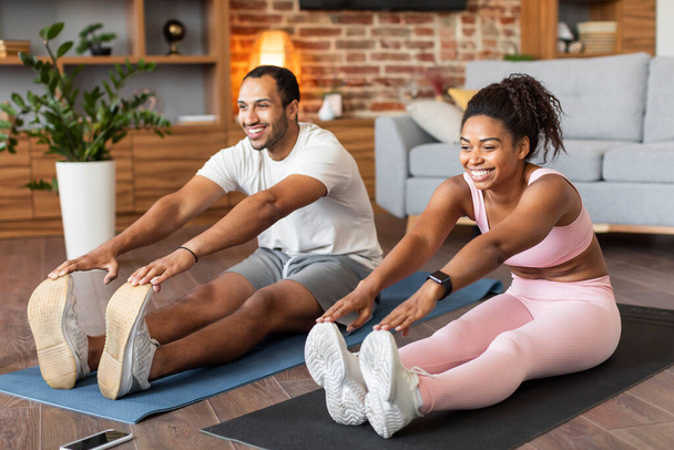 スポーツウェアで幸せな若い黒のカップルは、リビングルームのインテリアで一緒にトレーニング、床にマットの上に足のストレッチ演習を行います。健康管理,アクティブなライフスタイル,原因covid-19で自宅で痩身フィットネス - 写真・画像