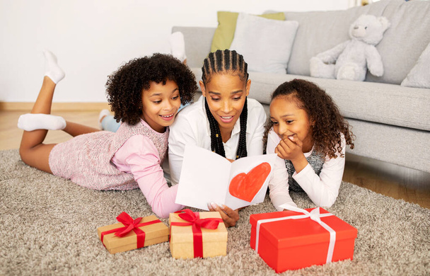 Glimlachende Afro-Amerikaanse moeder lezen wenskaart ontvangen geschenken van kleuters dochters op moederdag vakantie liggend op de vloer thuis. Familie vieren mama 's verjaardag Concept - Foto, afbeelding